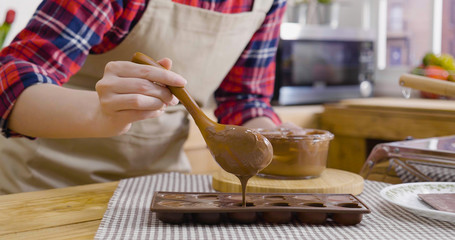 focus on female hand using wooden spoon pour filled in liquid cream melted dark chocolate heart love shape mold in modern kitchen at home. woman in apron doing handmade dessert process on valentines