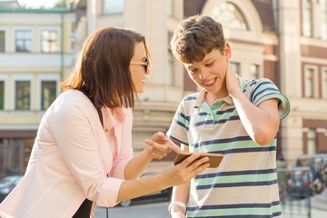 Parent and teenager, relationship. Mother shows her son something in mobile phone, boy is embarrassed, smiling, holding his hands on his head, city street background