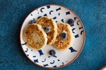 Cottage cheese pancakes on blue background. Syrniki with fresh berries and sour cream. Homemade food. Pancakes with cottage cheese on white plate with blueberry,