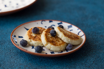 Cottage cheese pancakes on blue background. Syrniki with fresh berries and sour cream. Homemade food. Pancakes with cottage cheese on white plate with blueberry,