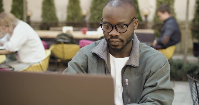 Smart afroamerican student wears eyeglasses working on laptop in outdoors cafe