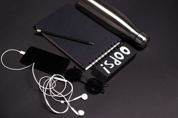 Top view of black work desk. Big eraser, glasses, pencil over notebook near smartphone with earphones and ecologic steel thermo bottle for water