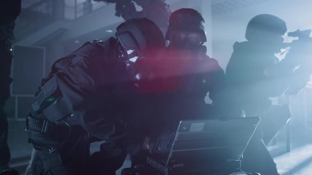 Masked Team of Armed SWAT Police Officers with Rifles are in Dark Seized Office Building with Desks and Computers. Soldier Opens a Laptop Computer to Plan a Tactical Attack.