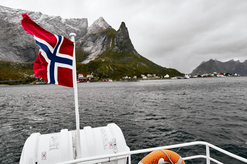 Norwegian flag in the wind on a boat deck while driving on the ocean with mountains and a village...