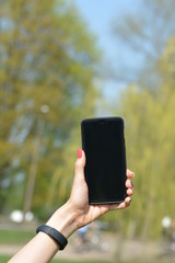 Woman's hand holding a smart-phone. Spring trees, blooming flowers and people in background. Soft focus, blur and bokeh