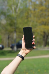 Woman's hand holding a smart-phone. Spring trees, blooming flowers and people in background. Soft focus, blur and bokeh