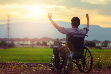 Close up of a paralyed man sitting on wheel chair raise hands up to the sky at rice green field with blurred village and mountain background in the evening 