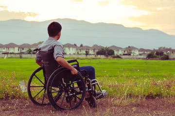 Close up of a paralyed young man sitting on wheel chair looking to rice green field with blurred village and mountain background in the evening 