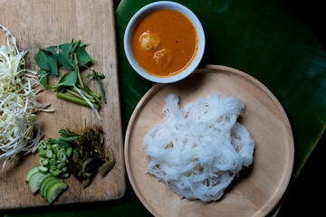 Rice noodles in fish curry sauce with vegetables on a wooden plate and place on the banana leaf floor. Separate coconut milk curry with meat ball and vegetables.
