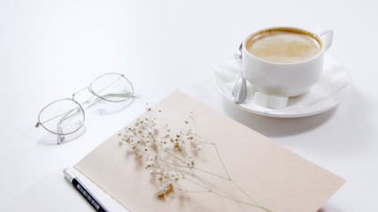On a white table there is a cup of coffee, glasses, a notebook with a pencil and a white flower.