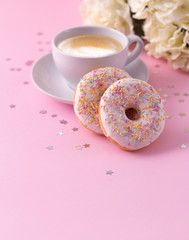 Mug of hot cappuccino and two donuts with flowers on a pink background.