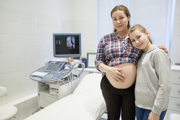 Twelve years old daughter standing with her pregnant mother in medical room with ultrasonic apparatus, unborn baby imaging on screen