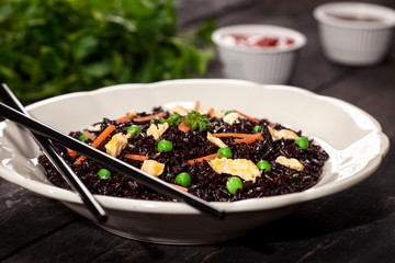 Cooked black rice with vegetables in a plate with sauces and chopsticks on the wooden background.