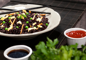 Cooked black rice with vegetables in a plate with sauces and chopsticks on the wooden background.