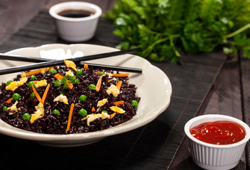 Cooked black rice with vegetables in a plate with sauces and chopsticks on the wooden background.