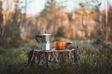 Coffee maker and wooden kuksa stand on a stump in the forest. Coffee maker and cup in focus. The...