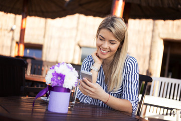 A beautiful young woman blonde is in a restaurant and she holds a smartphone in her hand and reads a message for which she is very happy.