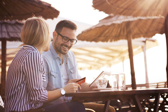 A handsome bearded young man and his pretty blonde girl sit in a restaurant on the seashore and they are happy because they bought something nice on the online shop.