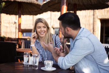 A handsome young man is jealous because his girlfriend talks too much over the phone while they drink coffee in a restaurant on the seashore.