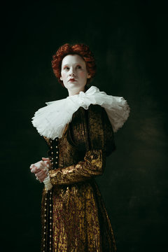 No fear for future. Medieval redhead young woman in golden vintage clothing as a duchess standing crossing hands on dark green background. Concept of comparison of eras, modernity and renaissance.