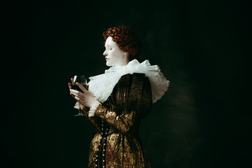 One part of me. Medieval redhead young woman in golden vintage clothing as a duchess holding a glass with red wine on dark green background. Concept of comparison of eras, modernity and renaissance.