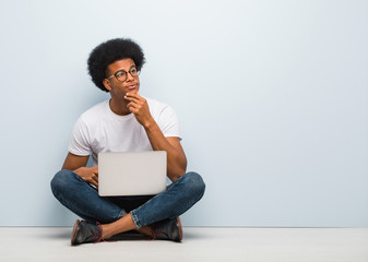 Young black man sitting on the floor with a laptop doubting and confused