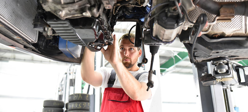 Automechaniker in Werkstatt repariert Fahrzeug // car mechanic in work clothes works in a workshop and repairs a vehicle