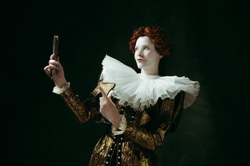 Medieval redhead young woman in golden vintage clothing as a duchess holding a mirror and a glass with white wine on dark green background. Concept of comparison of eras, modernity and renaissance.