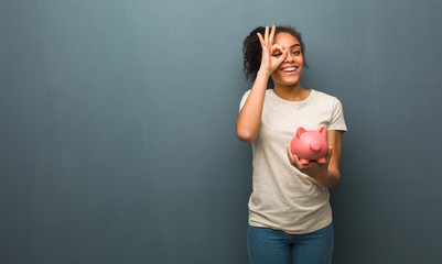 Young black woman confident doing ok gesture on eye. She is holding a piggy bank.