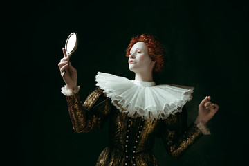 Proud of her self. Medieval redhead young woman in golden vintage clothing as a duchess looking in the mirror on dark green background. Concept of comparison of eras, modernity and renaissance. - obrazy, fototapety, plakaty