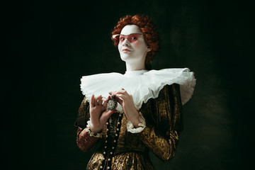 Secrets. Medieval redhead young woman in golden vintage clothing as a duchess in red sunglasses holding a parfume on dark green background. Concept of comparison of eras, modernity and renaissance.