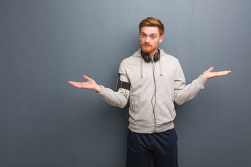 Young fitness redhead man doubting and shrugging shoulders