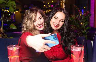 Women in red dresses drink cocktails and celebrate in a nightclub or bar. Friends take selfies and smile
