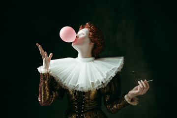 Medieval redhead young woman in golden vintage clothing as a duchess with red sunglasses blowing a bubblegum on dark green background. Concept of comparison of eras, modernity and renaissance.
