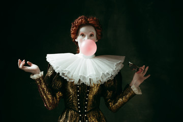 Medieval redhead young woman in golden vintage clothing as a duchess with red sunglasses blowing a bubblegum on dark green background. Concept of comparison of eras, modernity and renaissance.