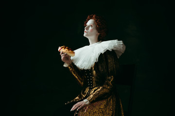 Dream about fit. Medieval redhead young woman in golden vintage clothing as a duchess eating a hot dog or sandwich on dark green background. Concept of comparison of eras, modernity and renaissance.