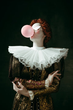 Medieval redhead young woman in golden vintage clothing as a duchess with red sunglasses blowing a bubblegum on dark green background. Concept of comparison of eras, modernity and renaissance.
