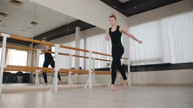 Graceful classical ballet dancer in sport clothes practicing standing split stretching position at ballet barre in dance studio.