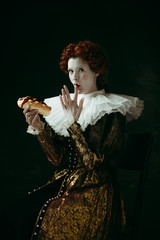 Only one bite. Medieval redhead young woman in golden vintage clothing as a duchess eating a hot dog or sandwich on dark green background. Concept of comparison of eras, modernity and renaissance.