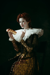 A little frolic. Medieval redhead young woman in golden vintage clothing as a duchess eating a hot dog or sandwich on dark green background. Concept of comparison of eras, modernity and renaissance.