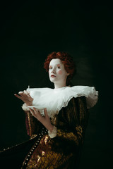 Medieval redhead young woman in golden vintage clothing as a duchess holding a cigarette and a cup of coffee on dark green background. Concept of comparison of eras, modernity and renaissance.