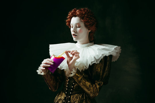 Taste of future. Medieval redhead young woman in golden vintage clothing as a duchess eating a fried potato on dark green background. Concept of comparison of eras, modernity and renaissance.
