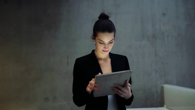 Young business woman with tablet walking in office, concrete wall in background.