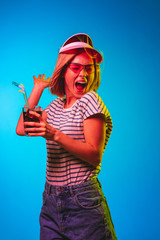 Beautiful female half-length portrait isolated on blue neon lights studio background. Young woman in red cap and sunglasses with drink. Facial expression, summer, weekend concept. Trendy colors.