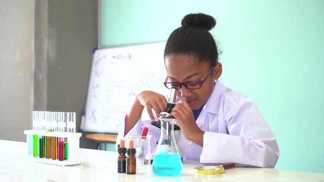Young African American kid using microscope and experimenting scientific lab along with chemical substance tubes and flasks in classroom - science and learning education concept