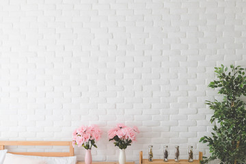 Dried flowers are packed in several clear glass bottles and pink rose on a wooden table in a vase beside the bed in the bedroom, with a white brick wall background.