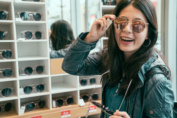 Smiling young asian woman traveler trying on sunglasses on mirror in optician face looking camera....