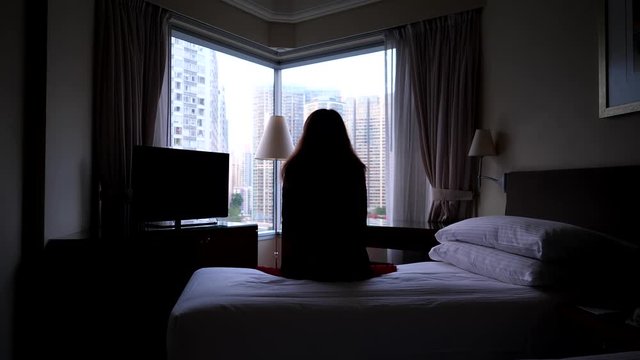 Sad woman in black suit and red skirt sit on bed, against corner window, looking outside. Lady stay still, dark room and bright city seen outdoors