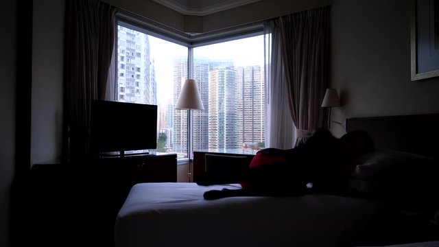 Woman feel sick, come and lay down on bed in suit and skirt, dark bedroom, bright city buildings seen outdoors through window glass. Long haired lady become unwell or sad, rest alone at dim room