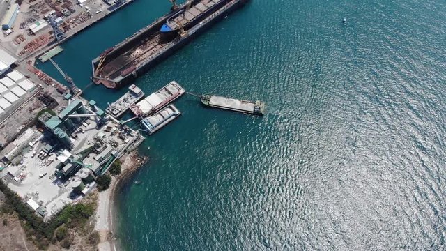 Dry cargo ships and floating dock facility near industrial shore, aerial shot. Small factory located on coast for easy loading, full vessels stay near pier. One ship stay under repair works